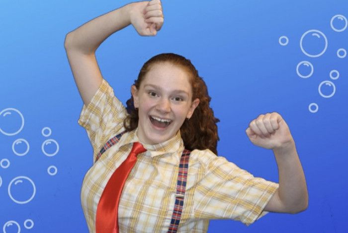 Girl in yellow checked shirt, red tie and suspenders, both fists in the air with an open-mouthed grin, against a blue background with bubbles