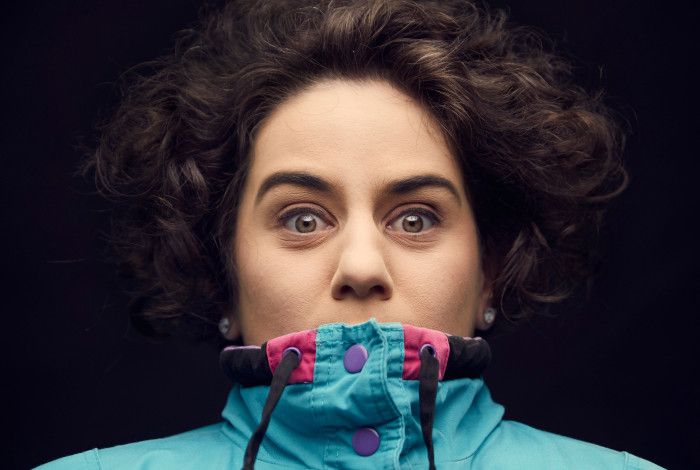 Jude Perl, against a black background, wearing a bright blue and pink parka pulled up over her mouth