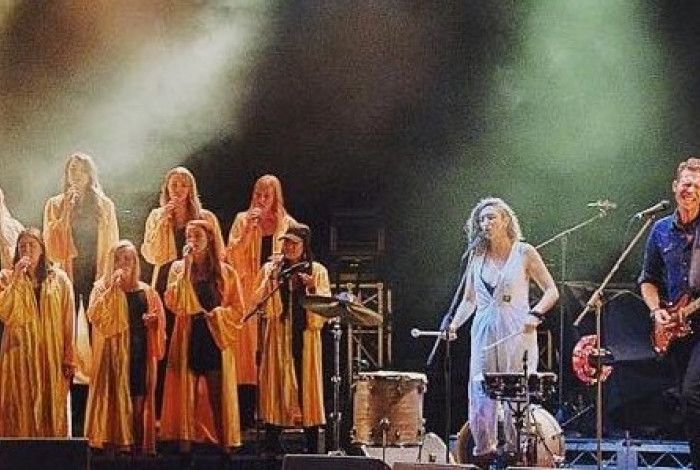 A choir dressed in yellow robes performing onstage, alongside a woman in a white dress and a man in a blue shirt. The man and woman are singing and the woman is playing drums, the man guitar
