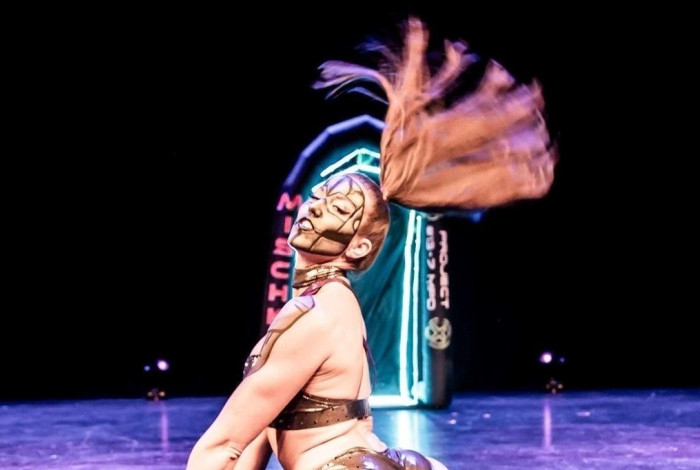 A pole dancer on a stage floor, flicking her large ponytail back, wearing metallic dancewear and face paint