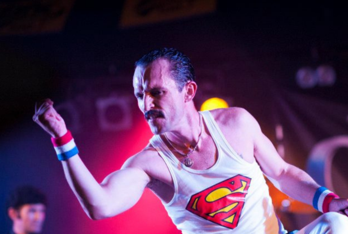 A performer dressed in white pants and white singlet with Superman emblem, representing Freddie Mercury of Queen
