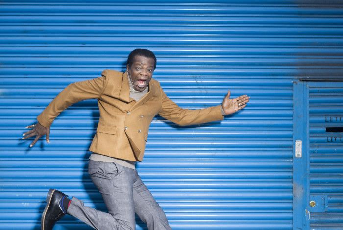 Comedian Stephen K Amos, jumping animatedly, against a bright blue corrugated iron backdrop