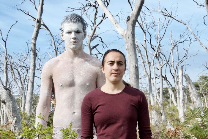 Two people standing in bushland, one is shirtless and covered in white body paint.
