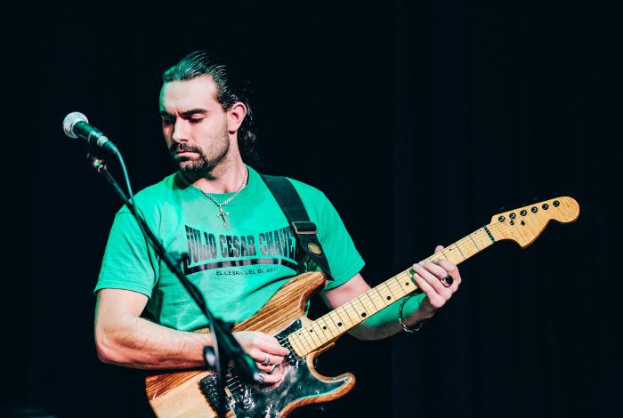 A man in a green t-shirt, with long dark hair in a ponytail, and facial hair, playing electric guitar onstage
