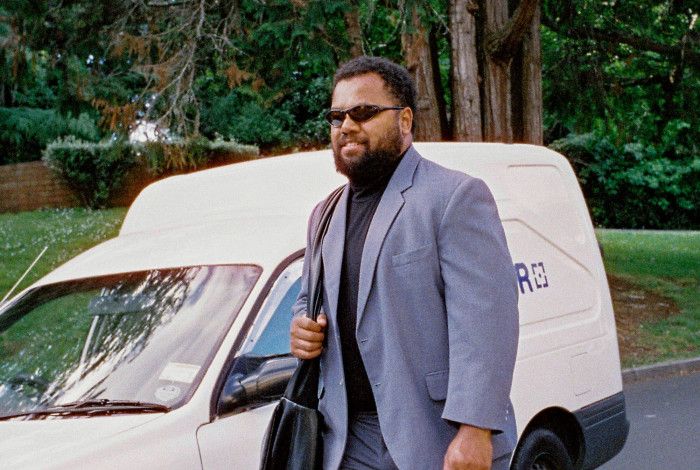 Man in light blue suit, black turtle neck and wrap around sunglasses carrying a messenger bag as he walks past a white delivery wagon