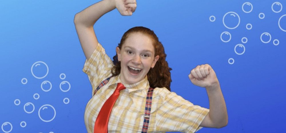 Girl in yellow checked shirt, red tie and suspenders, both fists in the air with an open-mouthed grin, against a blue background with bubbles