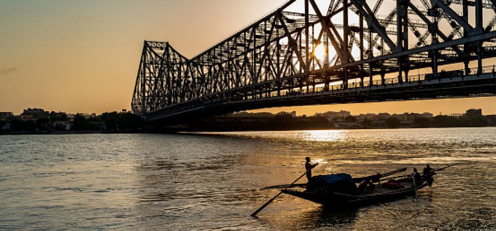 People in a small boat, silhouetted on water at sunrise or sunset, with a large bridge in the background