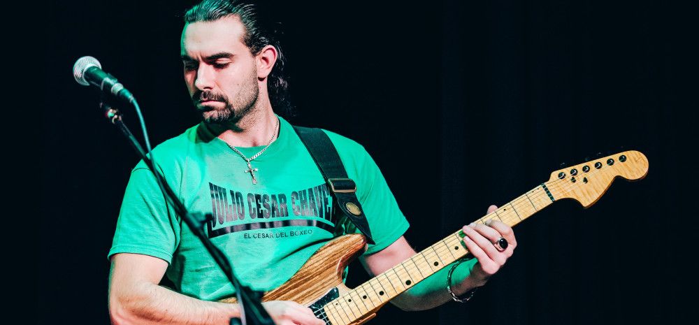 Simeon Boyadjiev, playing guitar, wearing a green tshirt, against a black stage background