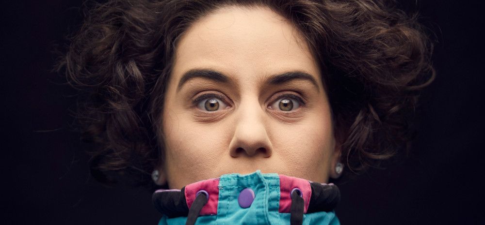 Jude Perl, against a black background, wearing a bright blue and pink parka pulled up over her mouth