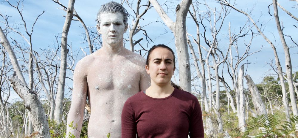 Two people standing in bushland, one is shirtless and covered in white body paint.