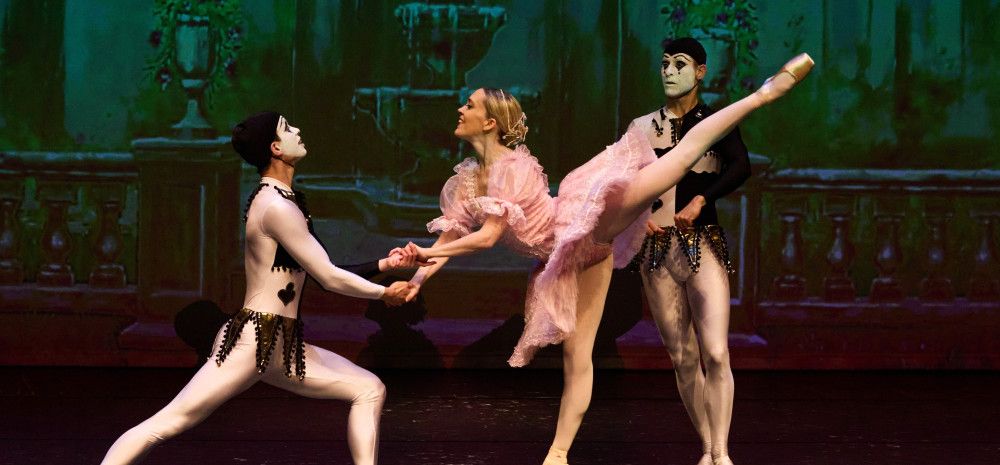 A female ballet dancer in pink costume dances with two male dancers dressed as harlequins