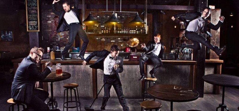 A bar setting, with five young men in tuxedos, dancing and striking various poses