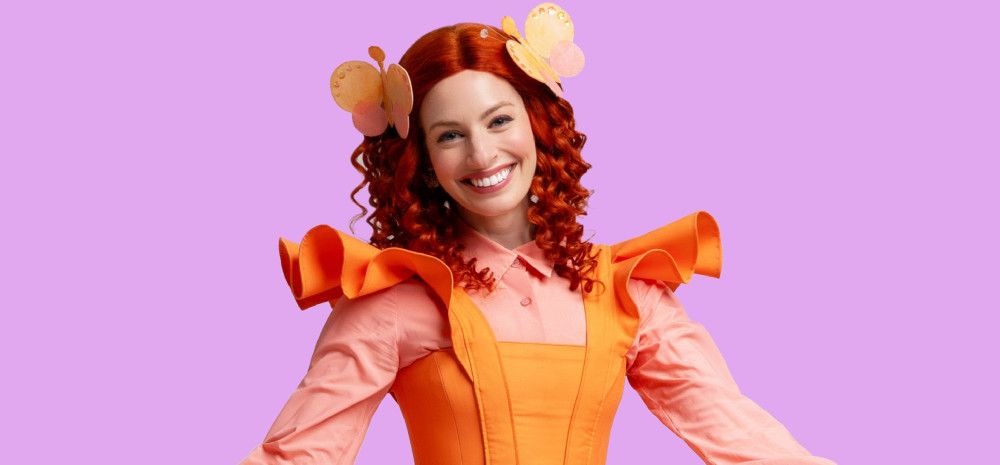 Emma Memma, dressed in a bright orange frilled pinafore dress and dusty pink shirt, tightly curled hair adorned with butterflies, smiling against a bright lavender background