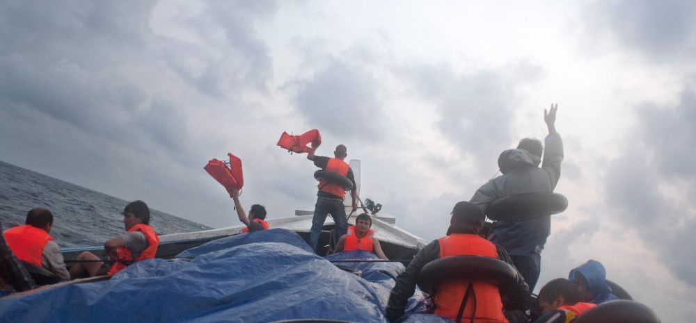 Figures on a boat, in life-vests, holding life vests aloft, against an overcast sky