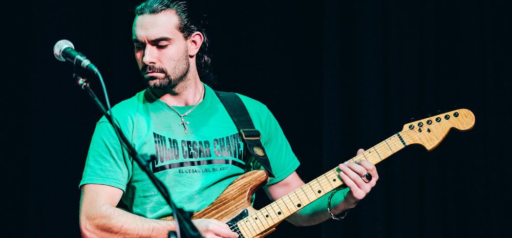 A man in a green t-shirt, with long dark hair in a ponytail, and facial hair, playing electric guitar onstage