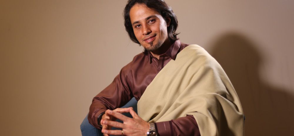 A man in a brown shirt, sitting with his fingertips resting together, looking peaceful, with a cream wrap over his shoulder