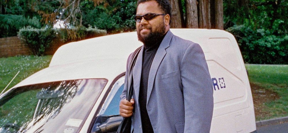 Man in light blue suit, black turtle neck and wrap around sunglasses carrying a messenger bag as he walks past a white delivery wagon